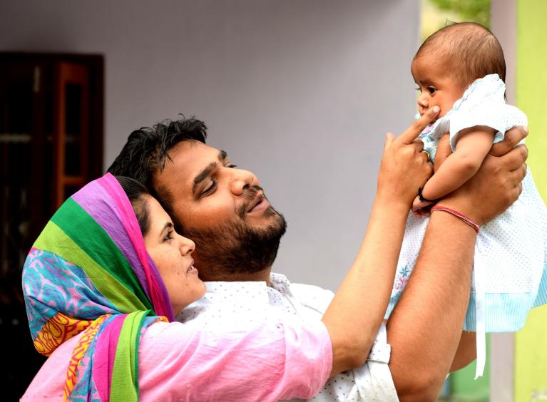 A parent holds their baby in the air whilst the child's other parent, touches their cheek with their finger.