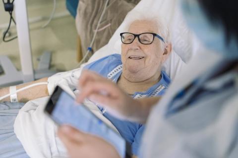 An older patient smiles in bed as a nurse inputs information on a tablet.