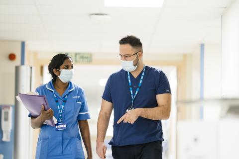 Two nurses wearing masks, talk as they walk down a corridor