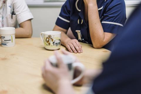 Close-up of a nurse holding a hot drink whilst sitting at a table, with two other staff partially shown in the background.