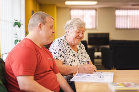 An person of "mid-age" looks on, as an older person smiles, whilst colouring in