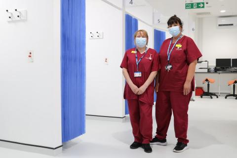 Two healthcare assistants in red scrubs, smile from behind masks, whilst standing in a clinical area of the CDC